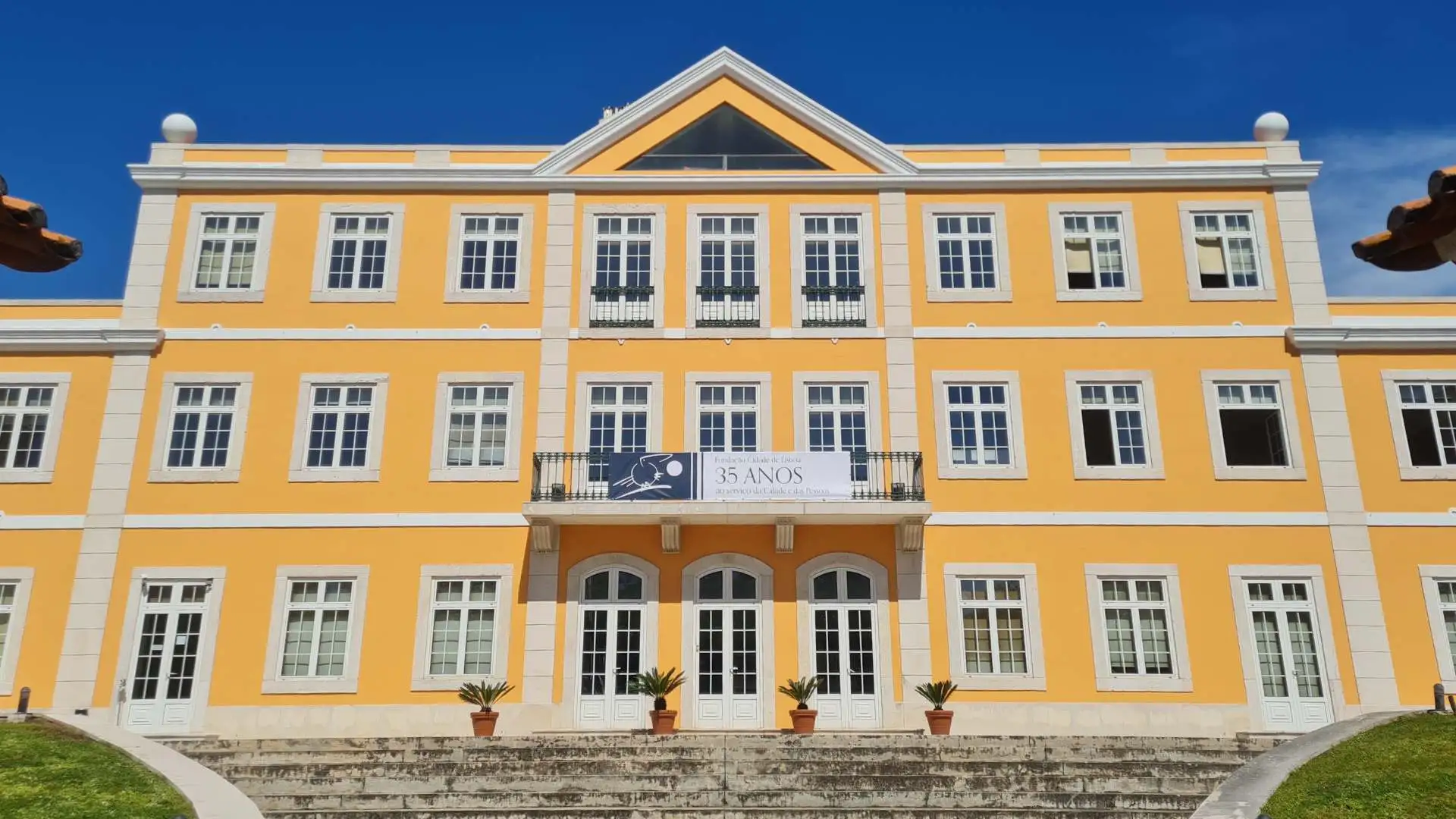 Fachada da sede da Fundação Cidade de Lisboa, edifício amarelo de três andares com estilo clássico e bandeira comemorativa dos 35 anos.